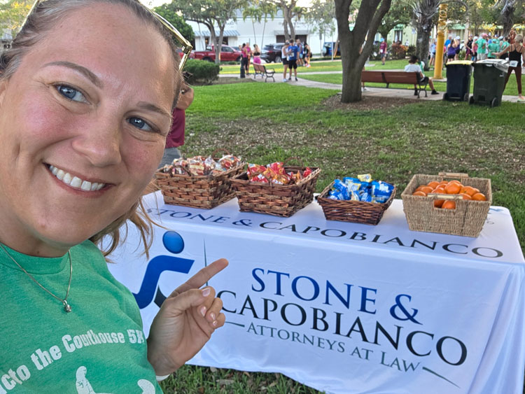 Smiling woman in a green shirt takes a selfie at an outdoor event next to a Stone & Capobianco Attorneys at Law table with baskets of snacks behind her.