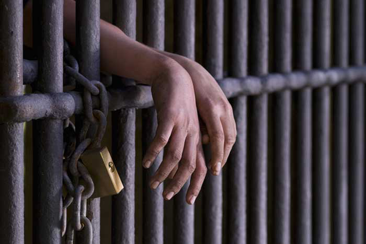 Hands resting over metal bars of a jail cell, with a chain and padlock visible on the bars.