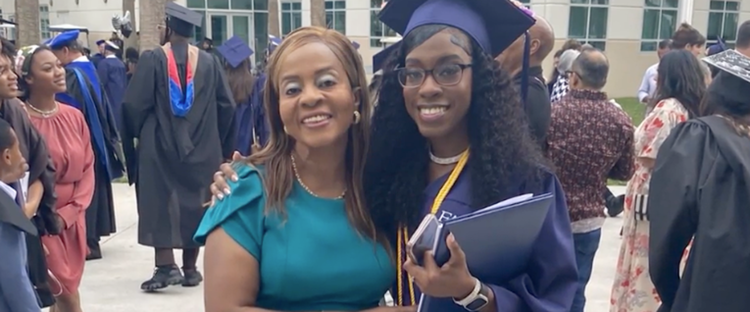 Marcia Pochette-Jenice Woods - Stone & Capobianco - Attorneys at Law Graduate in a purple cap and gown posing with a smiling woman in a teal dress at an outdoor graduation, holding folders.